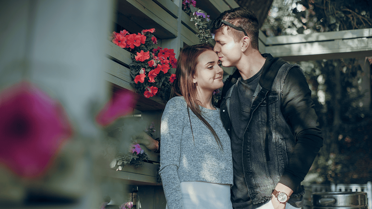 Zamilovaný pár při předsvatebním focení stojí u dřevěné pergoly s květinami. Přirozené focení snoubenců v romantickém prostředí plném barevných květů.